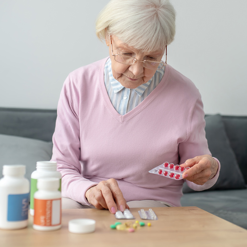 Eine ältere Frau sitzt auf der Couch in ihrem Zuhause und sortiert Medikamente auf einem Tisch.
