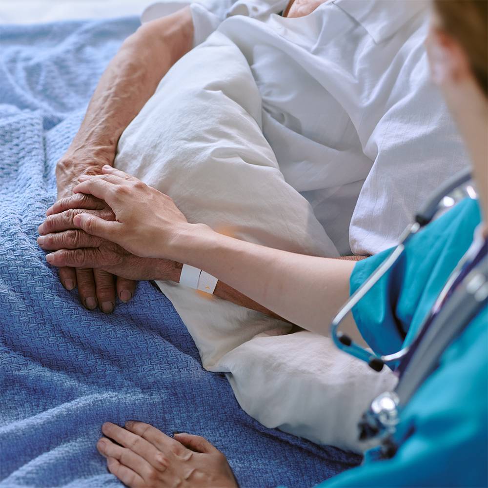 Detailansicht Palliativbetreuung. Die Hausärztin hält die Hand einer im Bett liegenden älteren Person und spricht mit ihr.