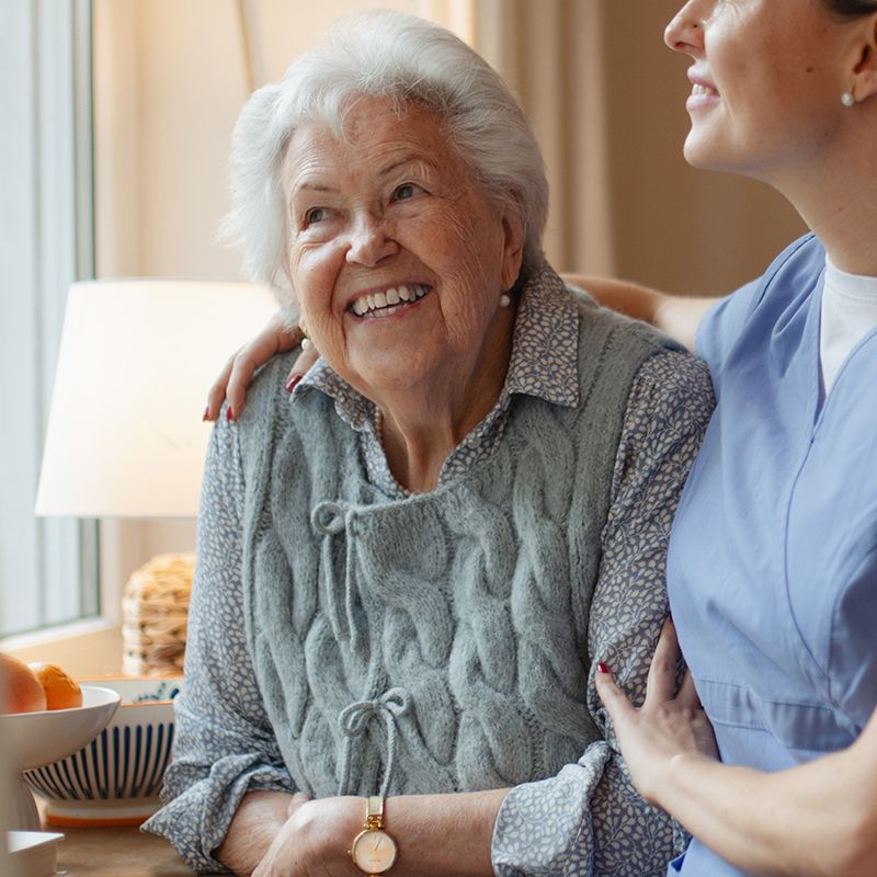 Eine alte Frau sitzt am Fenster und lächelt glücklich. Eine Arztfachkraft umarmt sie liebevoll.